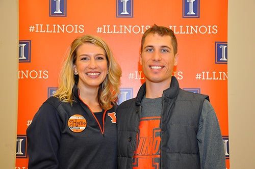 Two alumni wearing orange and blue pose in front of an Illini backdrop