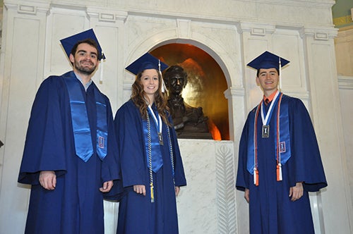 Three graduating scholars wearing caps and gowns stand next to the bust of Abraham Lincoln in the lobby of Lincoln Hall.